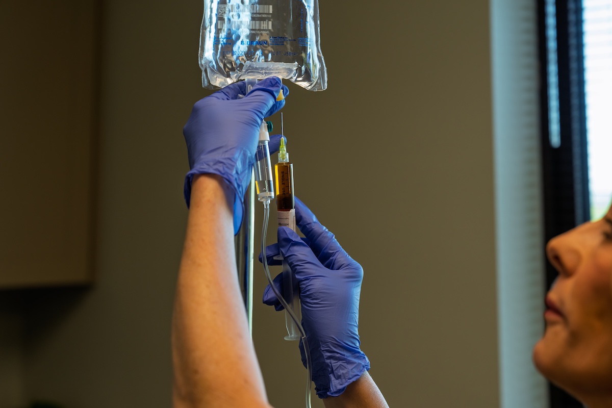 Gloved clinical staff hand preparing an IV nutrient bag and syringe for infusion